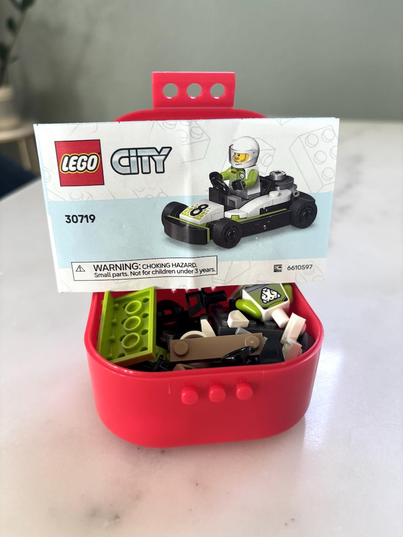 Puede incluir: Un contenedor de almacenamiento LEGO City rojo con un coche de carreras parcialmente ensamblado y una hoja de instrucciones impresa. La hoja muestra un coche de carreras con un conductor. El contenedor est&aacute; lleno de varias piezas de LEGO.