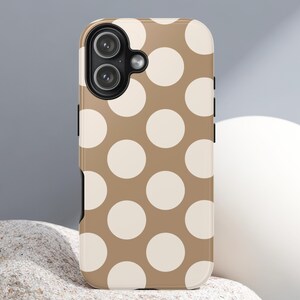 May include: A phone case with a tan background and large white polka dots. The case has a glossy finish and a black camera module. The phone case is designed to fit a smartphone.