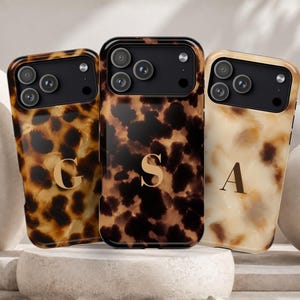 May include: Three smartphone cases featuring a tortoiseshell pattern in varying shades of brown, black, and cream. Each case has a black camera cutout and a gold-colored letter monogram in the center, displaying the letters G, S, and A respectively.