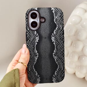 May include: A smartphone case with a snakeskin pattern. The case is black and white with a textured design. The case has a camera cutout and is held by a hand.