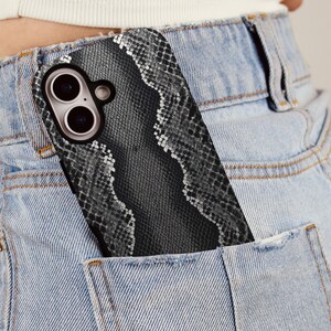 May include: A smartphone case with a snakeskin pattern is partially visible in the pocket of light blue denim jeans. The case has a black border and a gray and white snakeskin design. The phone's camera lenses are visible.