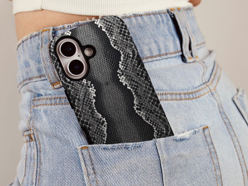 May include: A smartphone case with a snakeskin pattern is partially visible in the pocket of light blue denim jeans. The case has a black border and a gray and white snakeskin design. The phone's camera lenses are visible.