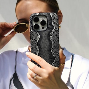 May include: A black and gray snakeskin-patterned phone case. The case has a textured appearance and a dark border. The phone case is held in front of a person wearing a white shirt and gold-rimmed sunglasses.