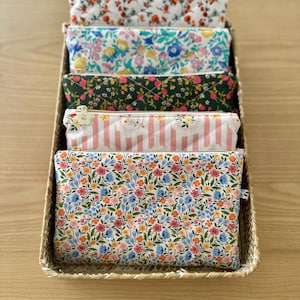 May include: A wicker basket contains six fabric pouches with floral patterns. The pouches display designs of red, blue, and pink flowers on white, and pink and white stripes. Each pouch has a zip closure. The pouches are made of fabric.