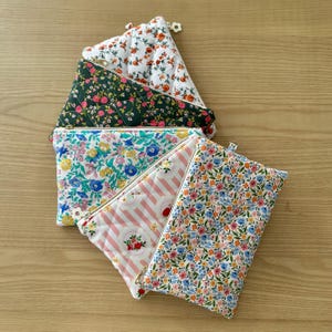 May include: Several quilted fabric pouches with floral and striped designs. The pouches display a range of colours, including white, pink, green, and blue, with gold zips. The pouches are arranged on a light brown wooden surface.