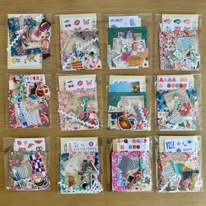May include: Twelve clear plastic bags, each packed with colourful paper ephemera, stickers, and fabric scraps. The bags contain a variety of decorative elements, including floral patterns, food illustrations, and text labels. They are arranged on a light brown wooden surface.