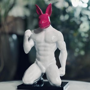 May include: A white sculpture of a kneeling figure with a muscular physique. The figure's head is a magenta rabbit mask. The sculpture is mounted on a black rectangular base, and the background is blurred.