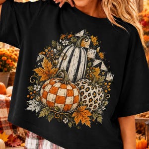 Peut inclure: Un t-shirt noir à manches courtes avec une impression graphique de trois citrouilles entourées de feuilles d'automne et de baies. La citrouille centrale a un motif rayé blanc et noir, une est à damier orange et blanc, et une est léopard.