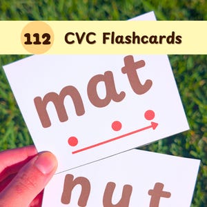 以下が含まれることがあります： 「mat」と「nut」と書かれた茶色の文字の白いフラッシュカード。 「mat」の単語には、赤い線と矢印が付いた点があります。 画像の上部には、黄色の背景に茶色の文字で「112 CVC Flashcards」というテキストがあります。