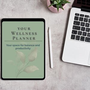 Puede incluir: Un planificador de bienestar digital en una tableta con el texto "YOUR WELLNESS PLANNER" y "Your space for balance and productivity". También se ven un lápiz óptico blanco, un ordenador portátil y una pequeña planta en maceta.