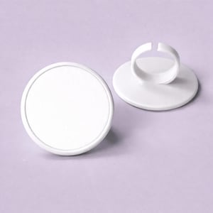 May include: Two white ring display stands. One stand is a flat, round disc. The other has a ring-shaped holder attached to a similar disc. Both are on a light purple background. These stands are ideal for showcasing rings.