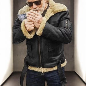 Può includere: Giacca bomber in montone nero con colletto e polsini in montone beige. La giacca ha una chiusura a zip, due tasche frontali e una cintura in vita. La persona nell'immagine indossa occhiali da sole e jeans blu.