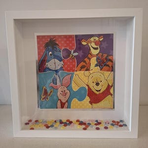May include: A framed artwork featuring a four-piece puzzle with beloved characters. Eeyore, Tigger, Piglet, and Winnie the Pooh are depicted in a colorful, dotted style. The white frame has a decorative bottom with colorful, small, round embellishments.