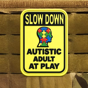 May include: A yellow patch with black trim and text that reads "SLOW DOWN" above "AUTISTIC ADULT AT PLAY." The patch features a silhouette of a head filled with puzzle pieces in various colors.