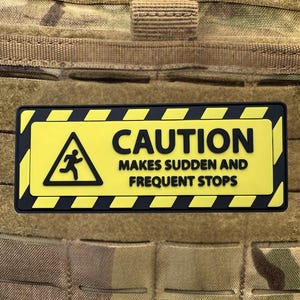 May include: A rectangular patch with a yellow background and black text that reads "CAUTION MAKES SUDDEN AND FREQUENT STOPS." The patch has a black border and a warning symbol of a running person.