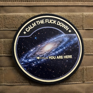 May include: A round patch with the text "CALM THE FUCK DOWN" in white on a black border. The center features a galaxy image with the words "YOU ARE HERE" and an arrow pointing to the galaxy.