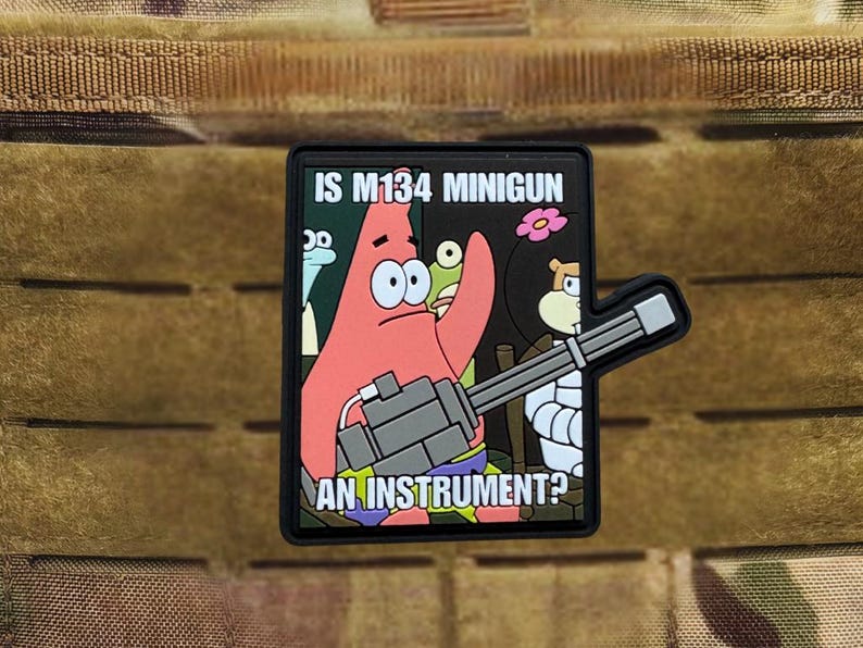 Puede incluir: Un parche negro con una imagen de dibujos animados de Patricio Estrella de Bob Esponja sosteniendo una minigun. El texto en el parche dice "IS M134 MINIGUN AN INSTRUMENT?" El parche tiene un respaldo de velcro.