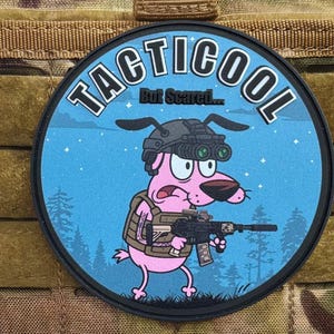 May include: Round patch with the text "TACTICOOL" and "But Scared...". The patch features a pink cartoon dog wearing night vision goggles and a tactical vest, holding a rifle. The background is blue with a forest silhouette.