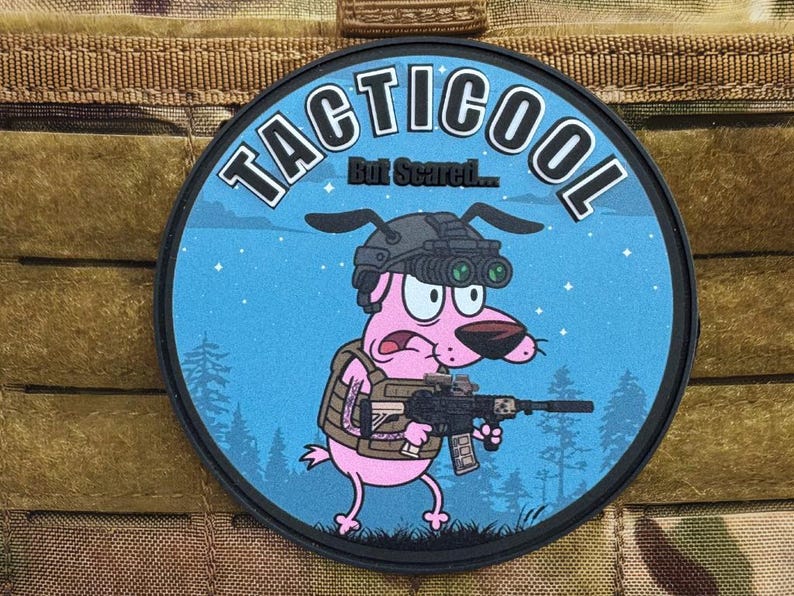 May include: Circular patch with the text "TACTICOOL" and "But Scared...". It depicts a pink cartoon dog in night vision goggles and a tactical vest, holding a rifle. The background is blue with a forest silhouette.