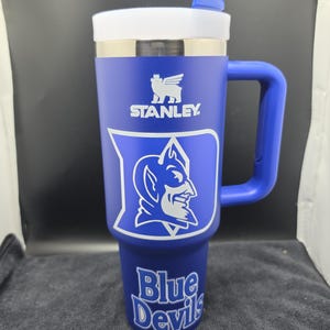 Pode incluir: Um copo Stanley azul com tampa e al&ccedil;a brancas. O copo apresenta o logotipo da Stanley e o logotipo Duke Blue Devils em branco. As palavras "Blue Devils" s&atilde;o impressas na parte inferior do copo.