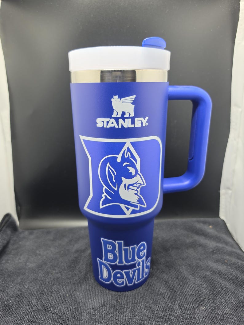 Pode incluir: Um copo Stanley azul com tampa e al&ccedil;a brancas. O copo apresenta o logotipo da Stanley e o logotipo Duke Blue Devils em branco. As palavras "Blue Devils" s&atilde;o impressas na parte inferior do copo.