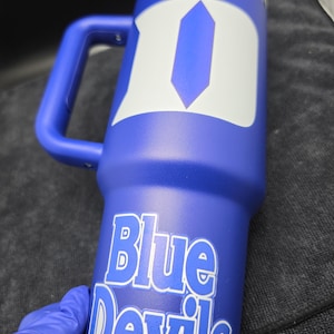 Pode incluir: Um copo t&eacute;rmico azul com uma al&ccedil;a e um design branco e azul. As palavras "Blue Devils" s&atilde;o impressas em branco no copo. O copo tem uma tampa prateada.