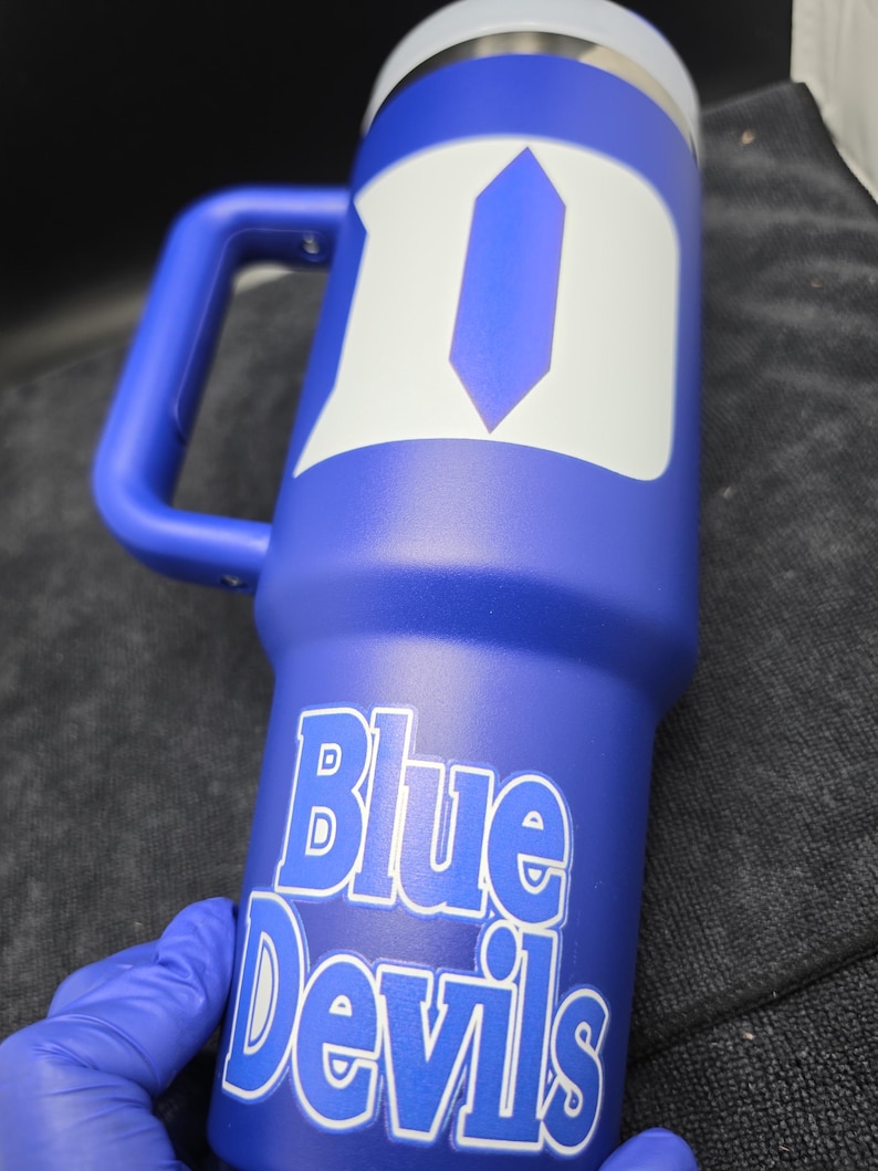 Pode incluir: Um copo t&eacute;rmico azul com uma al&ccedil;a e um design branco e azul. As palavras "Blue Devils" s&atilde;o impressas em branco no copo. O copo tem uma tampa prateada.