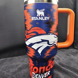 May include: A stainless steel Stanley travel mug with a blue, orange, and white Broncos design. The mug features the Broncos logo and the words "Bronco Denver". It has an orange handle and lid.
