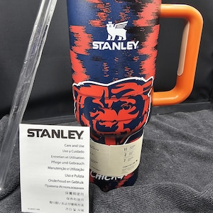 Op de afbeelding: Een Stanley reismok met een marineblauw en rood patroon, met het Chicago Bears logo. Het heeft een oranje handvat en een wit deksel. Inclusief een doorzichtig rietje en een verzorgingsinstructieblad.