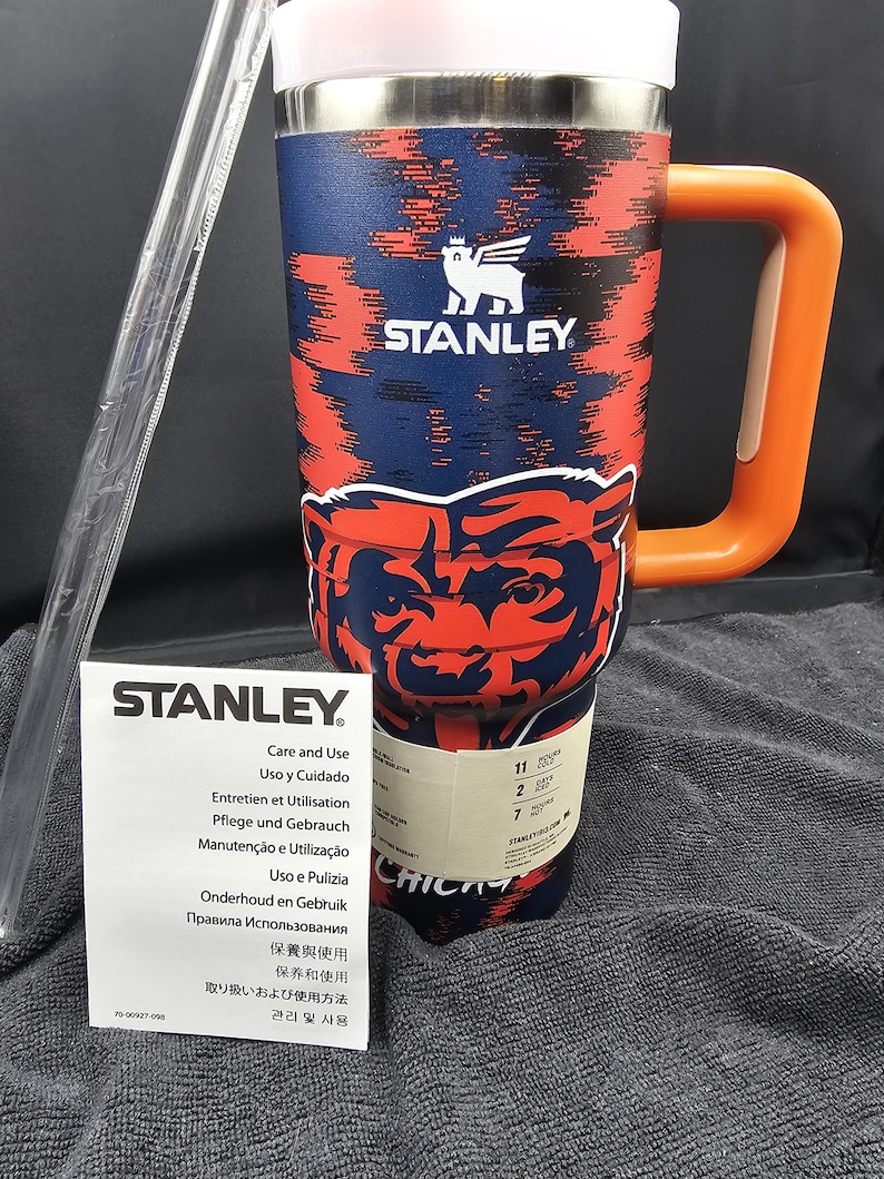 Op de afbeelding: Een Stanley reismok met een marineblauw en rood patroon, met het Chicago Bears logo. Het heeft een oranje handvat en een wit deksel. Inclusief een doorzichtig rietje en een verzorgingsinstructieblad.