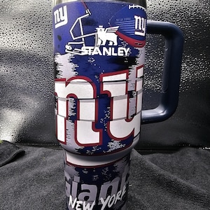 May include: A blue and white Stanley tumbler featuring the New York Giants logo. The tumbler has a large handle and a white lid. The design includes the team's logo, helmet, and the words "NY" and "Giants New York."
