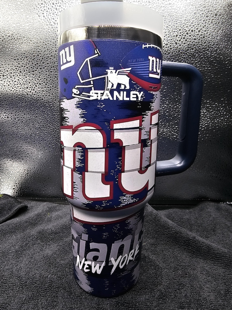 May include: A blue and white Stanley tumbler featuring the New York Giants logo. The tumbler has a large handle and a white lid. The design includes the team's logo, helmet, and the words "NY" and "Giants New York."