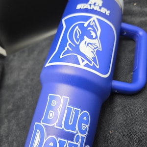 Pode incluir: Um copo Stanley azul com o logotipo branco de um diabo e as palavras "Blue Devils". O copo tem uma al&ccedil;a e uma tampa prateada. O logotipo da Stanley tamb&eacute;m &eacute; vis&iacute;vel.