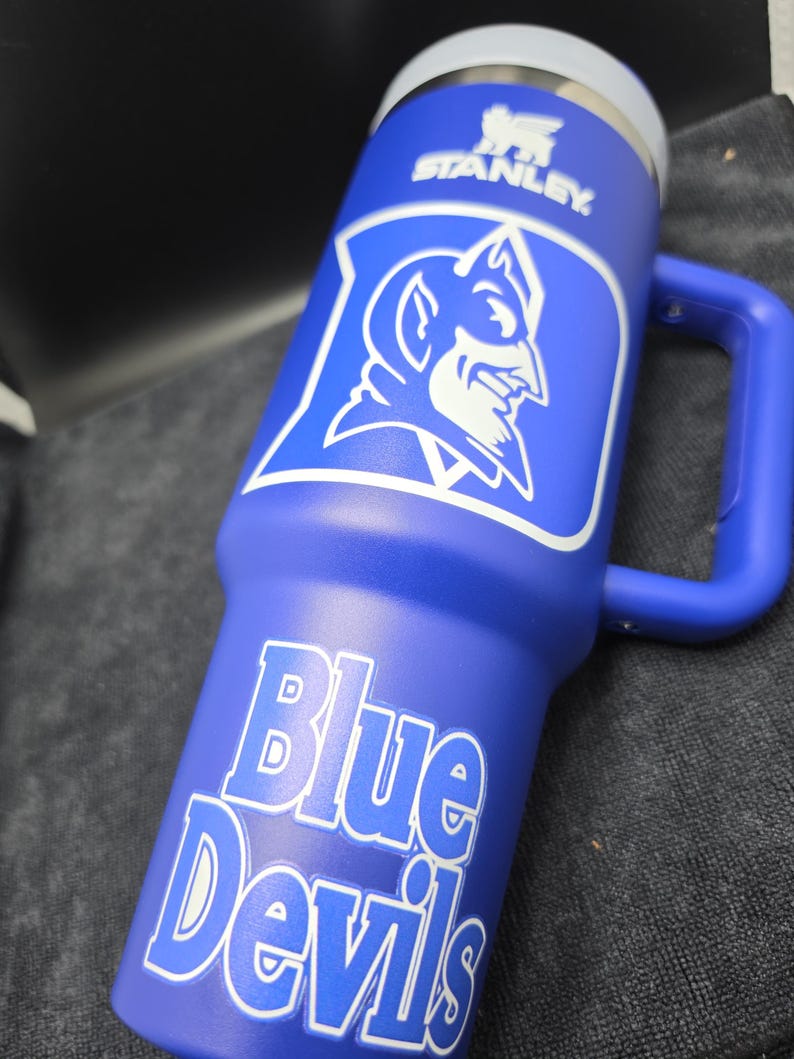 Pode incluir: Um copo Stanley azul com o logotipo branco de um diabo e as palavras "Blue Devils". O copo tem uma al&ccedil;a e uma tampa prateada. O logotipo da Stanley tamb&eacute;m &eacute; vis&iacute;vel.