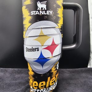 May include: A black and yellow Stanley travel mug featuring the Pittsburgh Steelers logo. The mug has a black handle and a lid. The Steelers logo is prominently displayed on the front, with the team name and city name also visible.