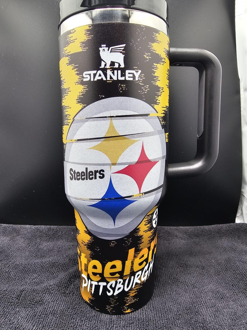 May include: A black and yellow Stanley travel mug featuring the Pittsburgh Steelers logo. The mug has a black handle and a lid. The Steelers logo is prominently displayed on the front, with the team name and city name also visible.