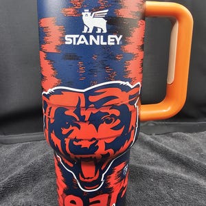 Op de afbeelding: Een roestvrijstalen Stanley-beker met een marineblauw en oranje ontwerp. De beker heeft het Chicago Bears-logo en het woord "Chicago". Hij heeft een oranje handvat en het Stanley-logo.
