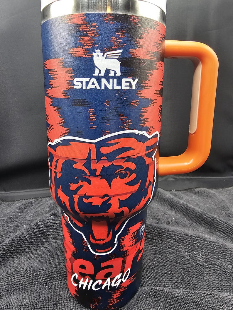Op de afbeelding: Een roestvrijstalen Stanley-beker met een marineblauw en oranje ontwerp. De beker heeft het Chicago Bears-logo en het woord "Chicago". Hij heeft een oranje handvat en het Stanley-logo.