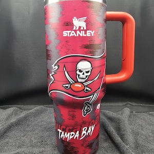 Puede incluir: Un vaso Stanley rojo y negro con tapa blanca y asa roja. El vaso presenta el logotipo de los Tampa Bay Buccaneers, una calavera con espadas cruzadas y un balón de fútbol, y las palabras "TAMPA BAY".