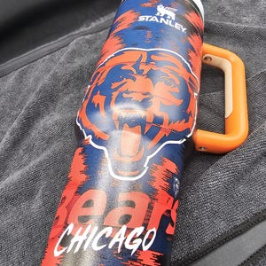Op de afbeelding: Een ge&iuml;soleerde Stanley-mok met een Chicago Bears-ontwerp. De mok heeft een blauw en oranje kleurenschema met het berenlogo van het team en het woord "Chicago" in het wit. Het heeft een oranje handvat en een witte rand.