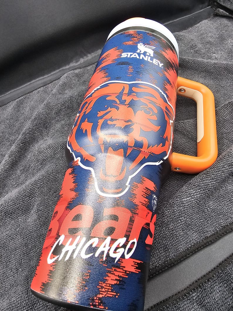 Op de afbeelding: Een ge&iuml;soleerde Stanley-mok met een Chicago Bears-ontwerp. De mok heeft een blauw en oranje kleurenschema met het berenlogo van het team en het woord "Chicago" in het wit. Het heeft een oranje handvat en een witte rand.