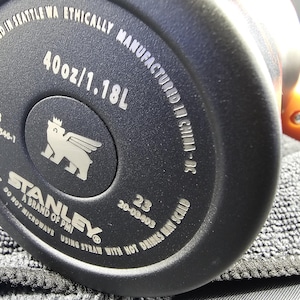 Op de afbeelding: Close-up van de bodem van een zwarte Stanley-beker. De tekst luidt "40oz/1.18L" en "Designed in Seattle WA Ethically Manufactured in China-DC". Het Stanley-logo is zichtbaar, samen met andere productinformatie.