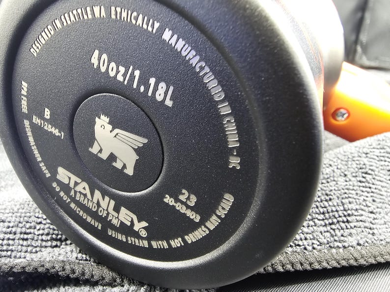 Op de afbeelding: Close-up van de bodem van een zwarte Stanley-beker. De tekst luidt "40oz/1.18L" en "Designed in Seattle WA Ethically Manufactured in China-DC". Het Stanley-logo is zichtbaar, samen met andere productinformatie.