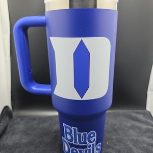 Pode incluir: Um copo t&eacute;rmico azul com tampa e al&ccedil;a brancas. O copo apresenta um grande logotipo "D" branco e as palavras "Blue Devils" em escrita branca. O copo foi projetado para conter aproximadamente 887 ml.
