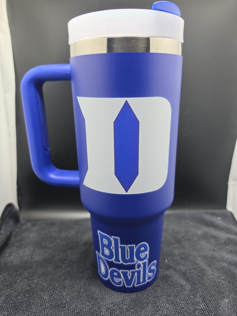 Pode incluir: Um copo t&eacute;rmico azul com tampa e al&ccedil;a brancas. O copo apresenta um grande logotipo "D" branco e as palavras "Blue Devils" em escrita branca. O copo foi projetado para conter aproximadamente 887 ml.