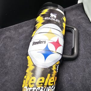 May include: A black and yellow Stanley travel mug featuring the Pittsburgh Steelers logo. The mug has a black handle and lid, with the team name and logo prominently displayed. The mug is designed for hot or cold beverages.