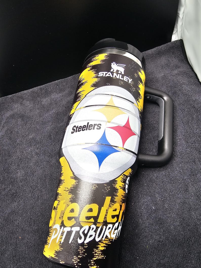 May include: A black and yellow Stanley travel mug featuring the Pittsburgh Steelers logo. The mug has a black handle and lid, with the team name and logo prominently displayed. The mug is designed for hot or cold beverages.