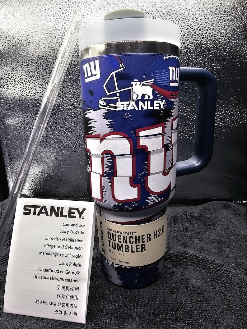 May include: A blue and white Stanley Quencher H2.0 tumbler with a handle, featuring the New York Giants logo. Includes a clear straw and a care and use booklet. The tumbler holds 1.18 litres.