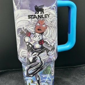 Puede incluir: Vaso Stanley de acero inoxidable con una ilustración de cómic de Tormenta, con cabello blanco y un traje plateado y negro. El vaso tiene un asa y tapa azules. La palabra "STORM" está impresa en la parte inferior.