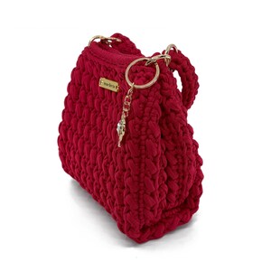 Può includere: Una borsa all'uncinetto rosso scuro con un motivo a maglia spesso e strutturato. La borsa presenta un'etichetta metallica dorata con il marchio Anna-Dorta e un portachiavi circolare dorato con un piccolo cristallo pendente sul manico.